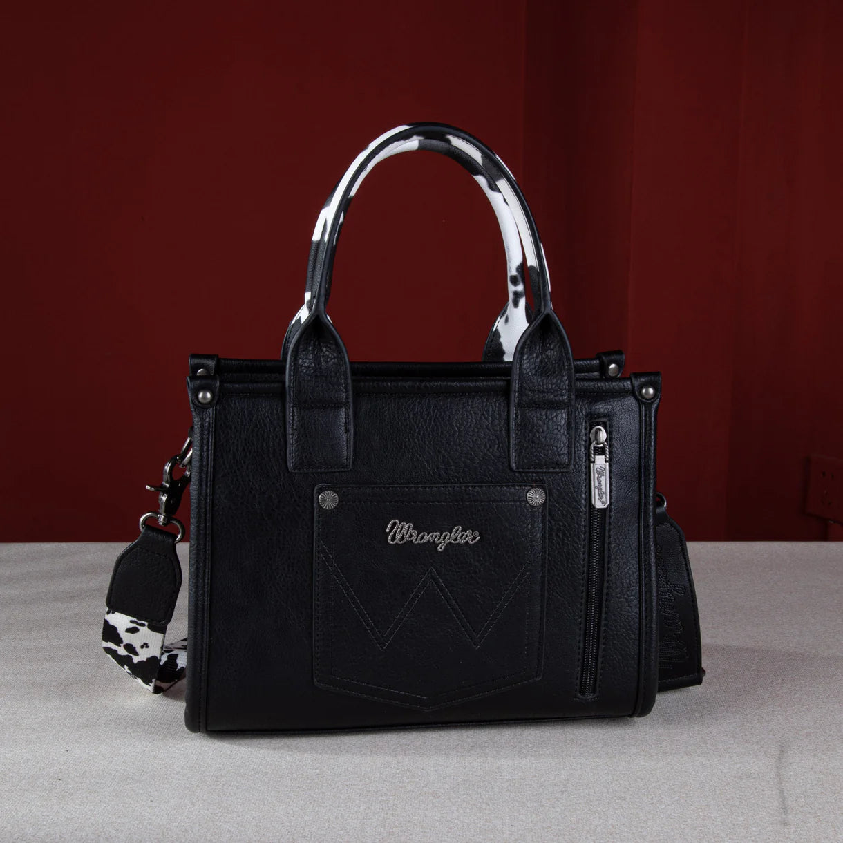 Front view: "Wrangler Cow Print Concealed Carry Tote Crossbody Bag Black — front view"
Interior/pocket view: "Wrangler Cow Print Concealed Carry Bag — interior concealed carry pocket detail"
Side/crossbody view: "Wrangler Black Cow Print Crossbody Bag — worn as crossbody style"