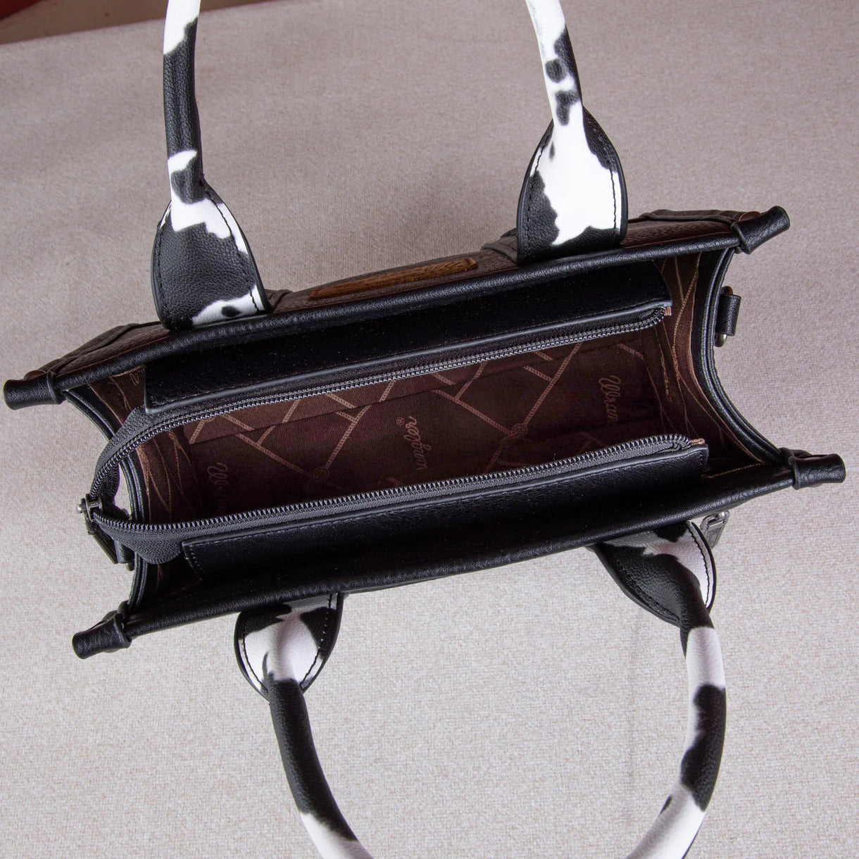Front view: "Wrangler Cow Print Concealed Carry Tote Crossbody Bag Black — front view"
Interior/pocket view: "Wrangler Cow Print Concealed Carry Bag — interior concealed carry pocket detail"
Side/crossbody view: "Wrangler Black Cow Print Crossbody Bag — worn as crossbody style"
